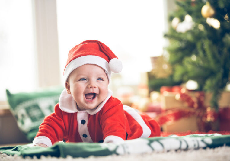 Cute little baby boy in Santa hat and clothes  lying down on the green blanket on the floor in front of the Christmas tree.